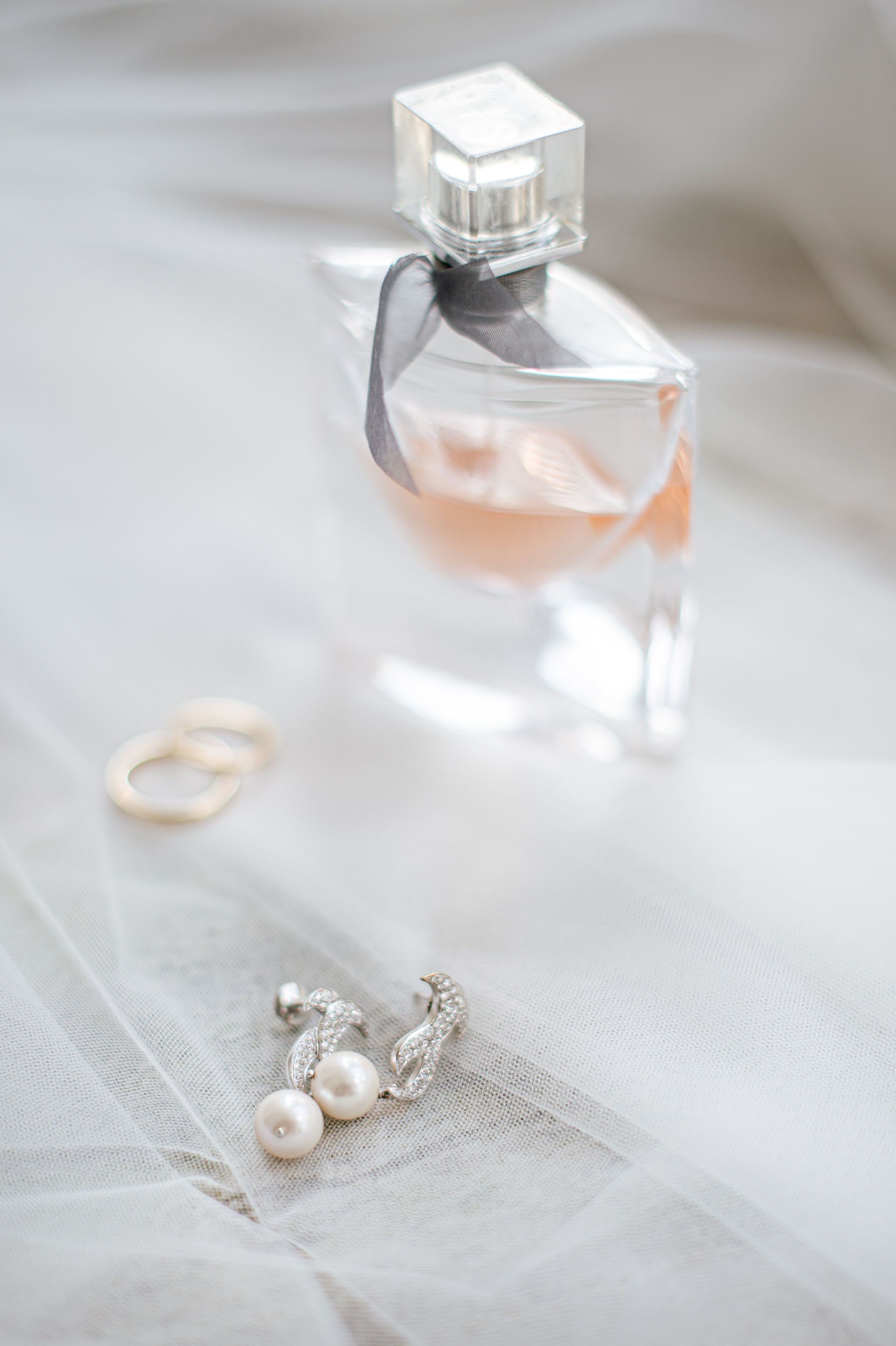 Perfume bottle and pearl earrings arranged on white fabric with soft lighting.