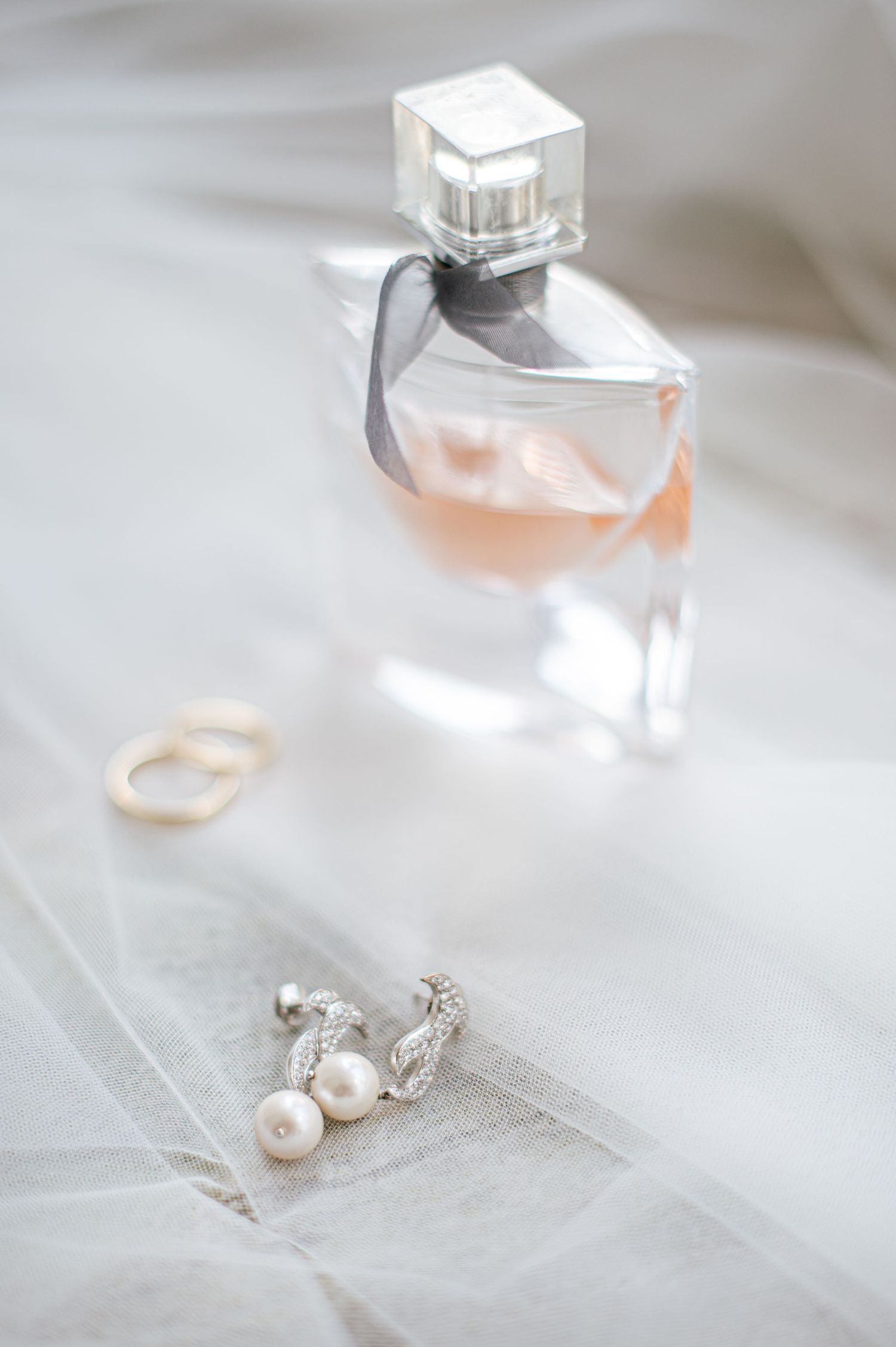 Perfume bottle and pearl earrings arranged on white fabric with soft lighting.