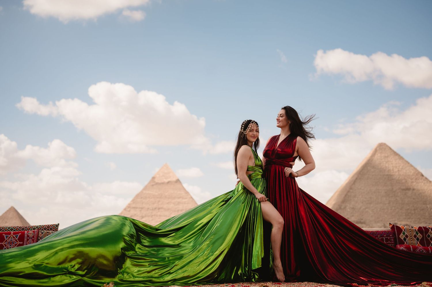 Emerald and burgundy dresses catch wind against pyramid landscape backdrop.