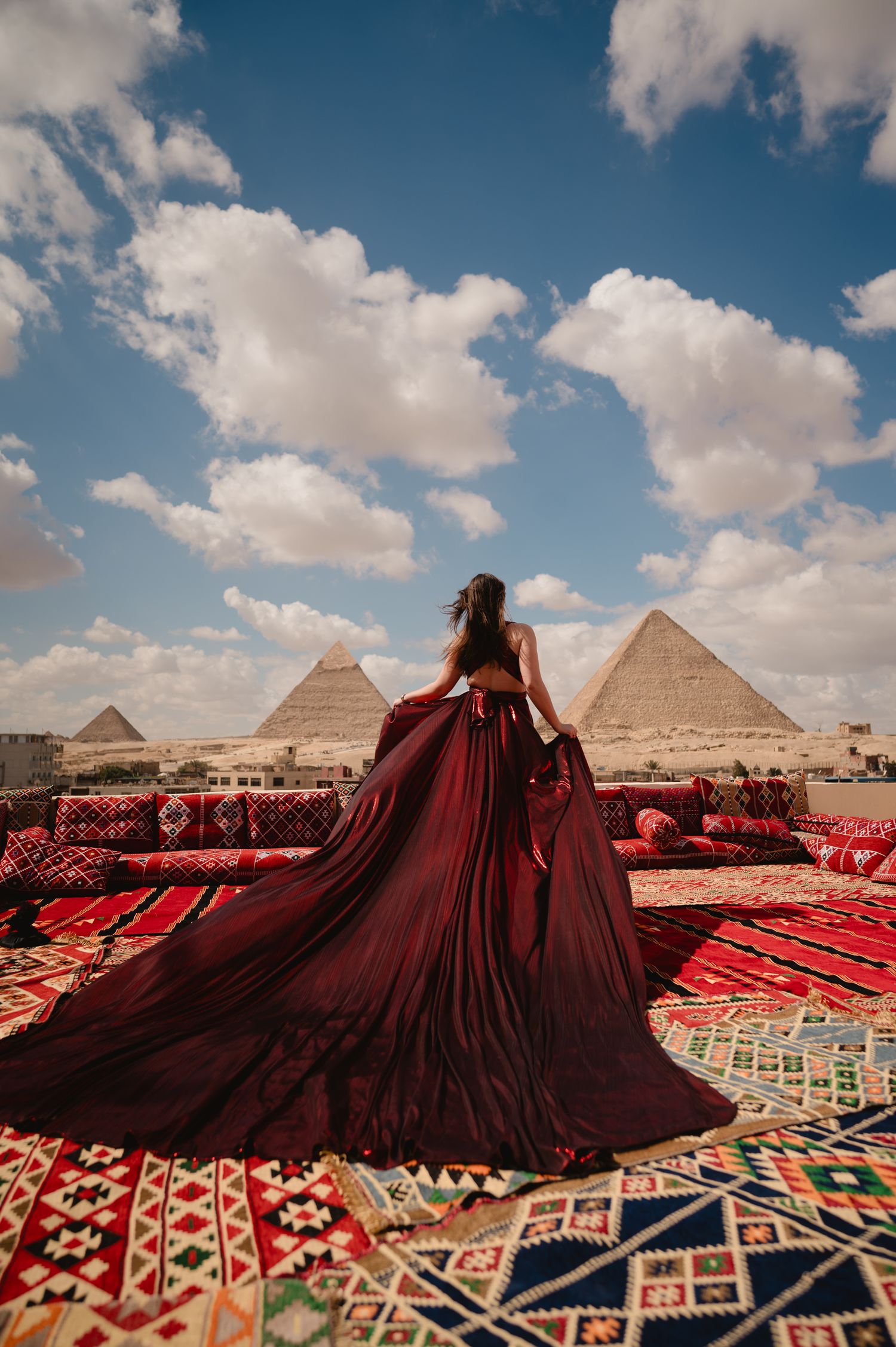 Flowing burgundy gown spreads across patterned carpets with pyramids in background.