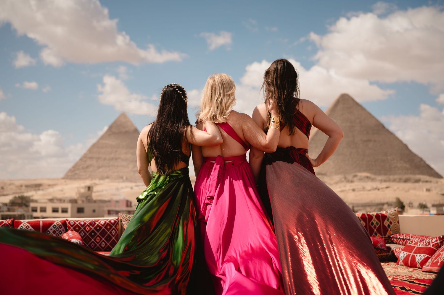 Three flowing dresses in green, pink and burgundy billow against the ancient Egyptian pyramids backdrop.