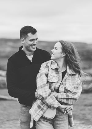 A candid outdoor portrait of a couple laughing together with windswept hair against a countryside backdrop.