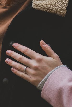 A close-up shot of a hand wearing a delicate engagement ring against a cozy sweater background.