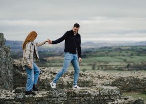 A couple holds hands while standing on elevated terrain with a scenic countryside view behind them.