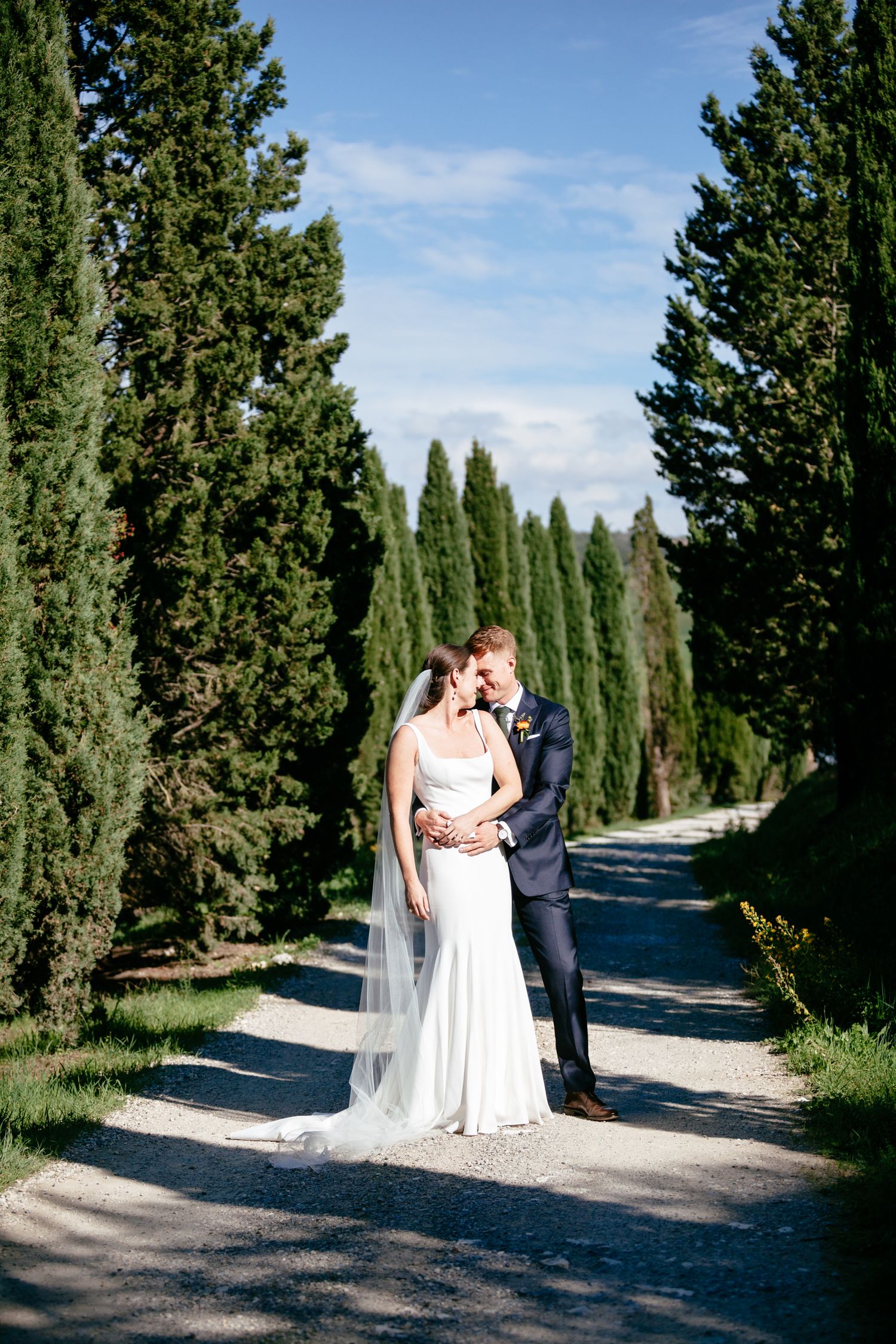 A bride and groom embrace along a tree lined avenue at the Lazy Olive in Tuscany.