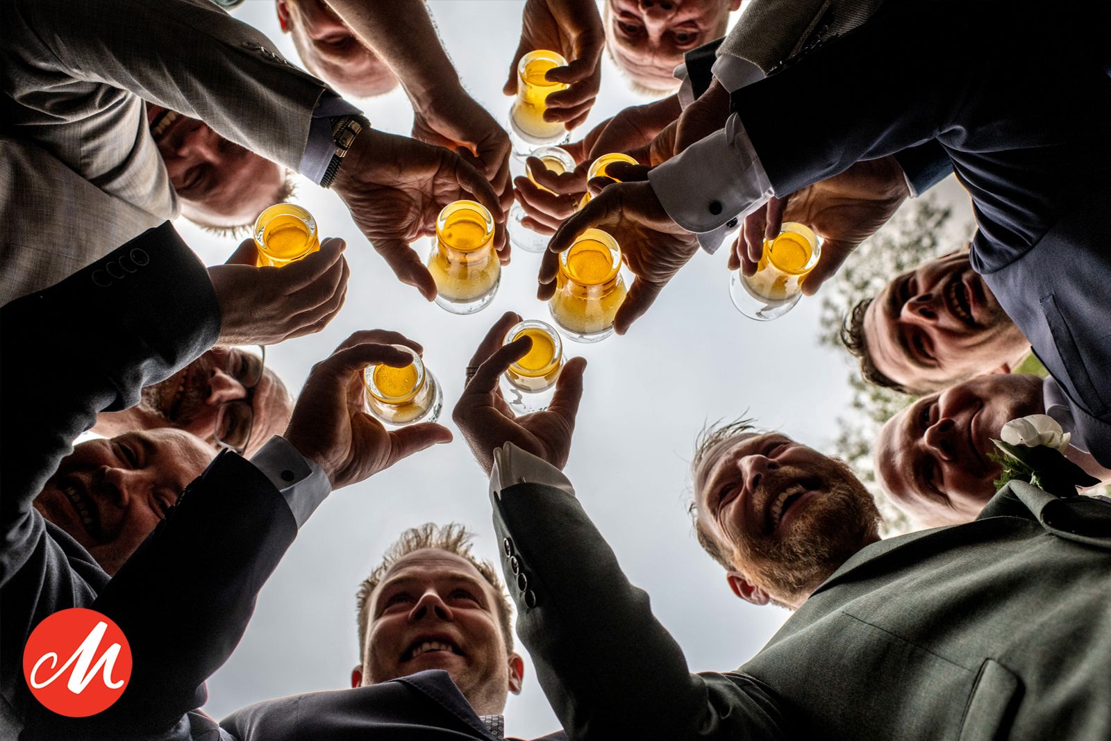 Friends toasting with beer glasses in a circular formation viewed from below against bright light.