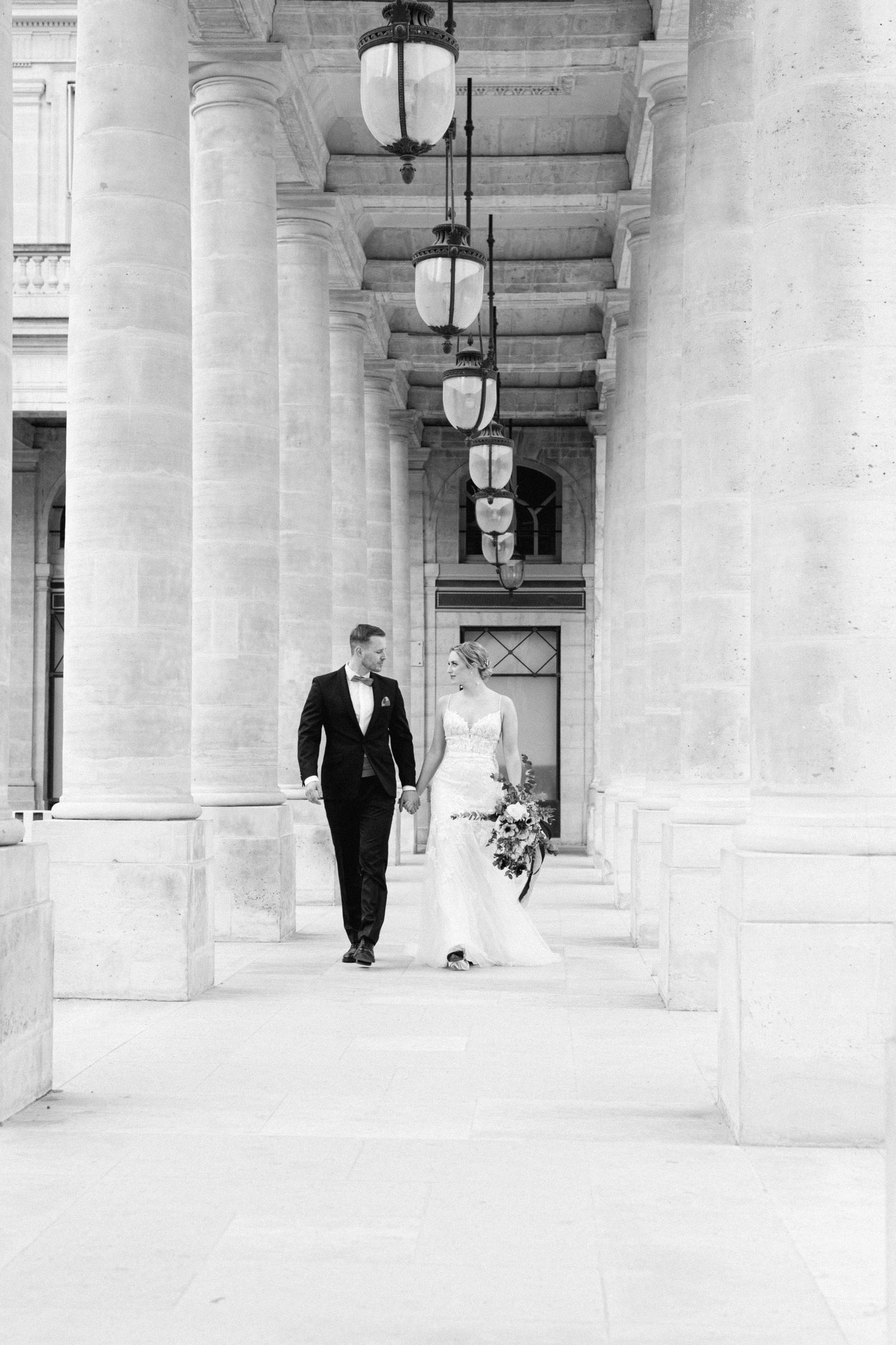 A couple walks through an elegant columned hallway with hanging lanterns in black and white photography in Paris.