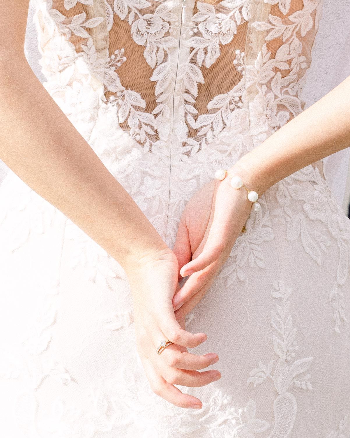 Close up view of delicate white lace details on a wedding dress with feminine hands clasped behind back.