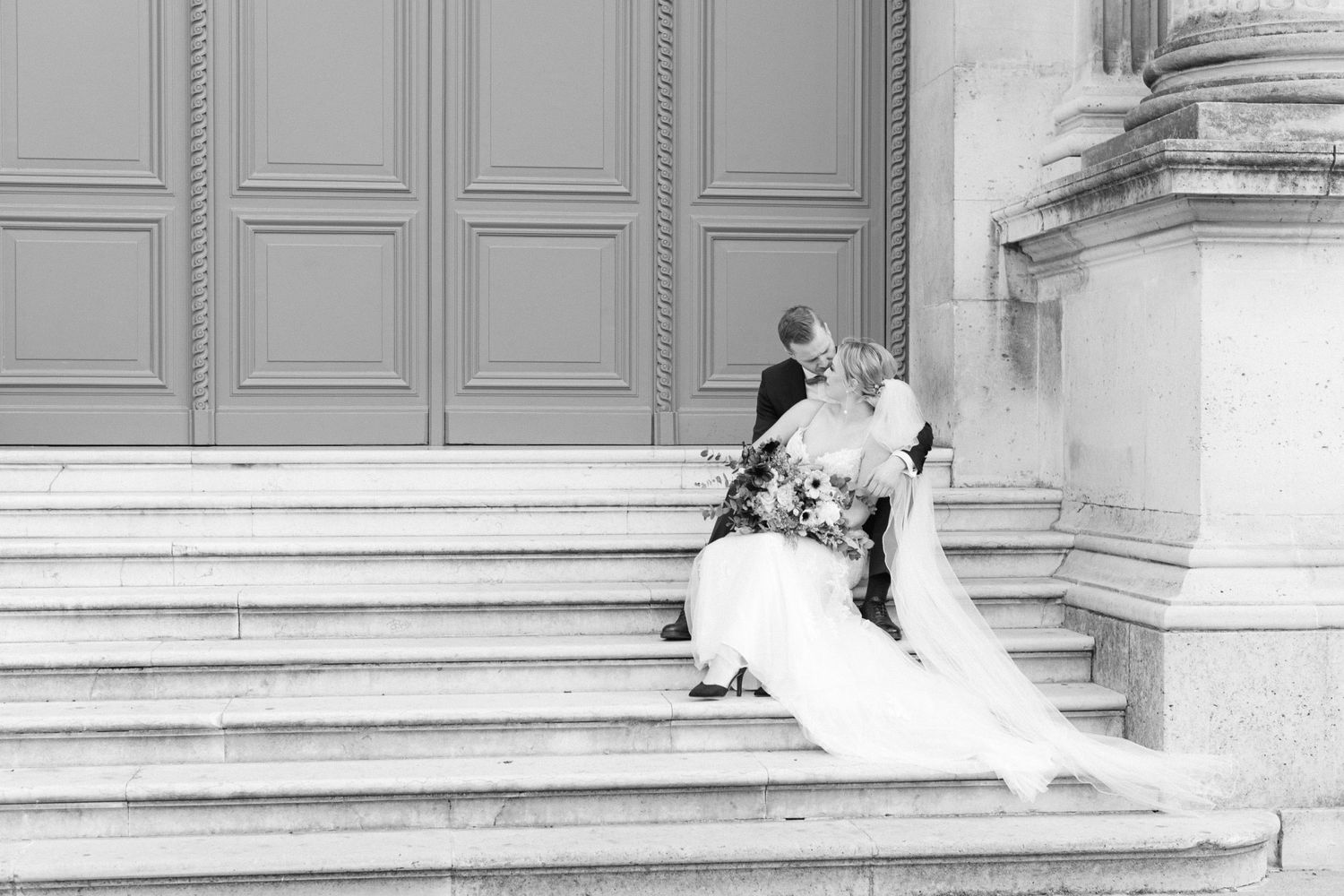 Black and white photo of a newlywed couple sitting on grand stone steps outside an elegant building in Paris.