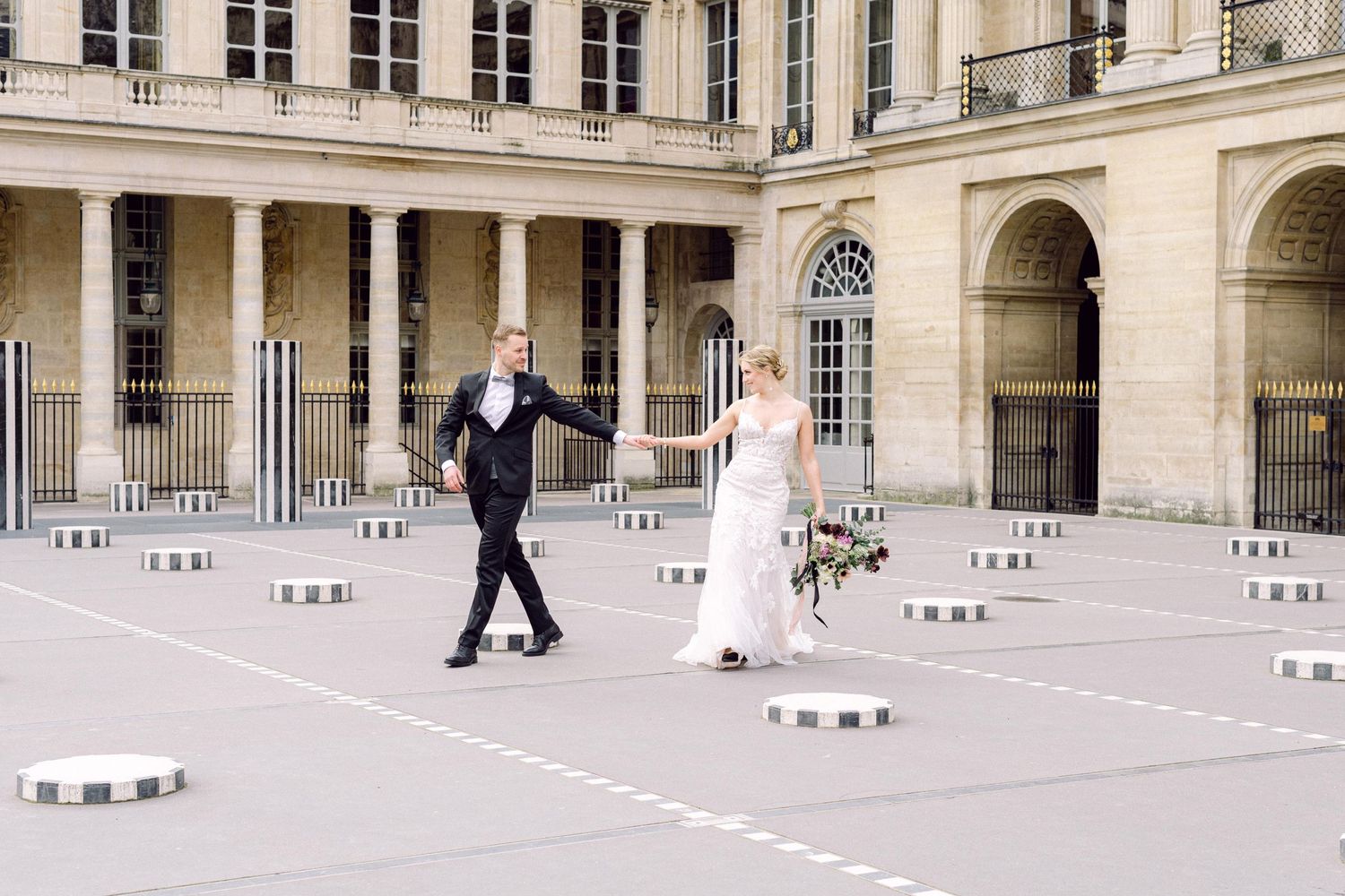 A couple walks together holding hands in an elegant Paris courtyard surrounded by classical architecture and stone pillars.