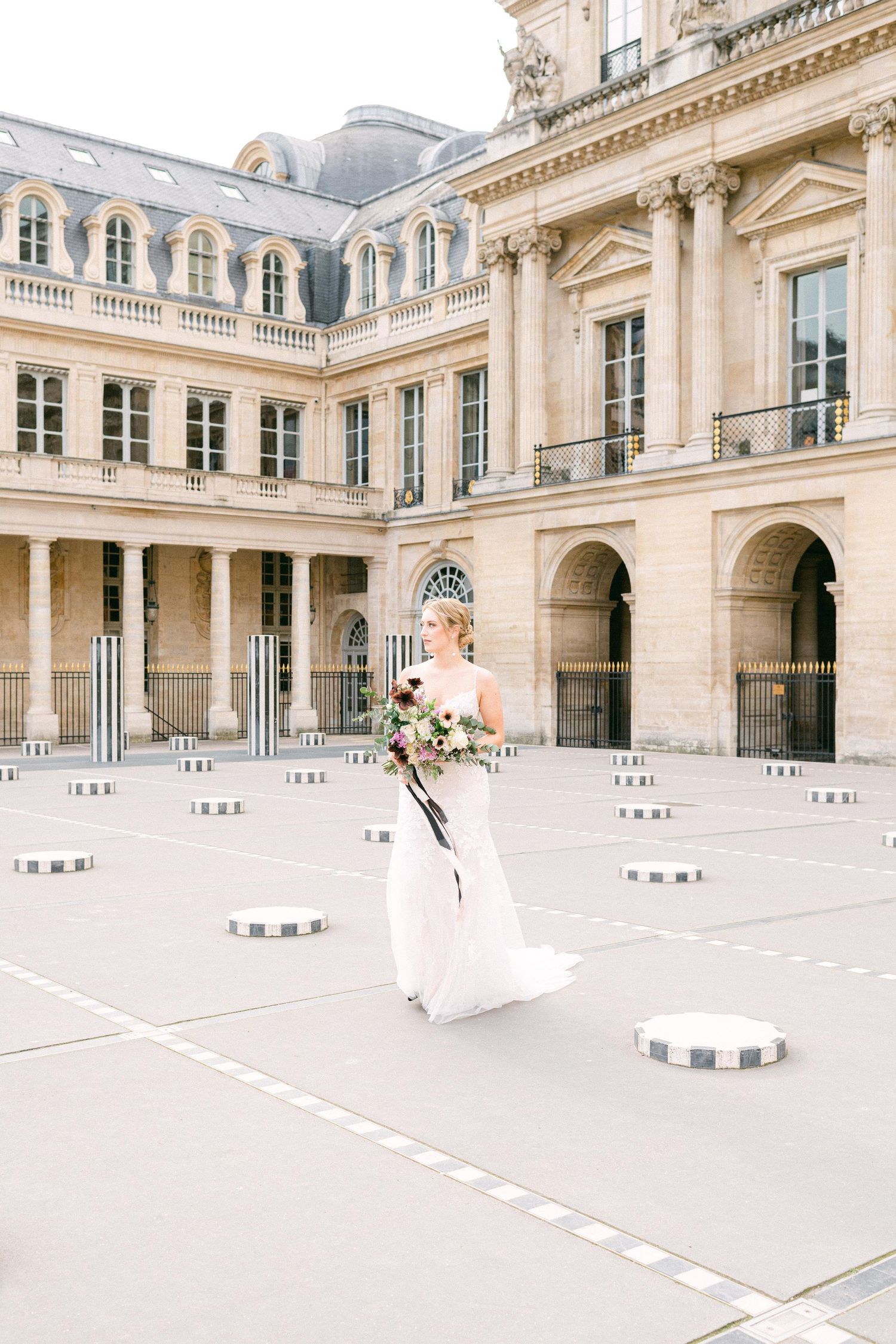 A bride holds a bouquet while standing in an elegant Paris palace courtyard with stone columns and architectural details.