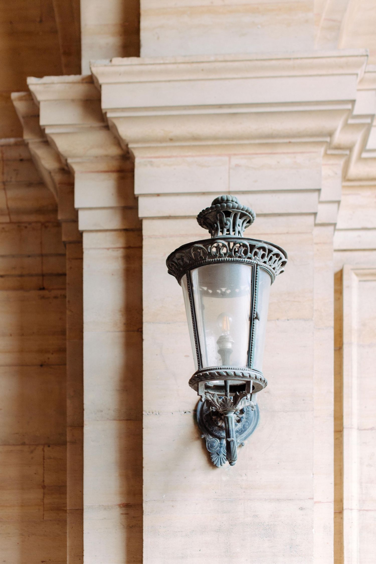 An ornate vintage wall-mounted lantern with decorative metalwork mounted on a beige stone pillar in a beautiful Paris yard.