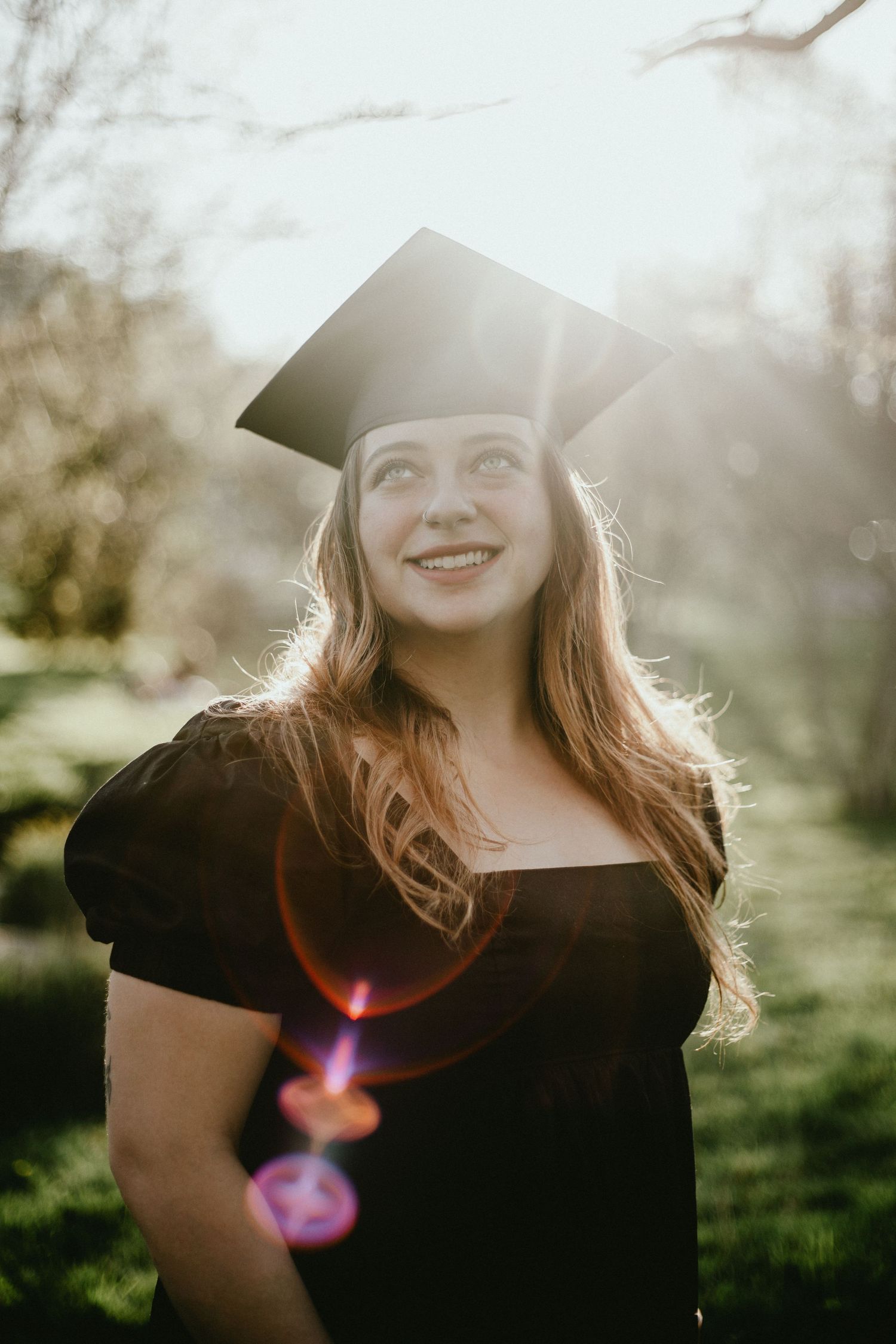 Graduate in black dress and cap smiling during golden hour outdoor photoshoot.