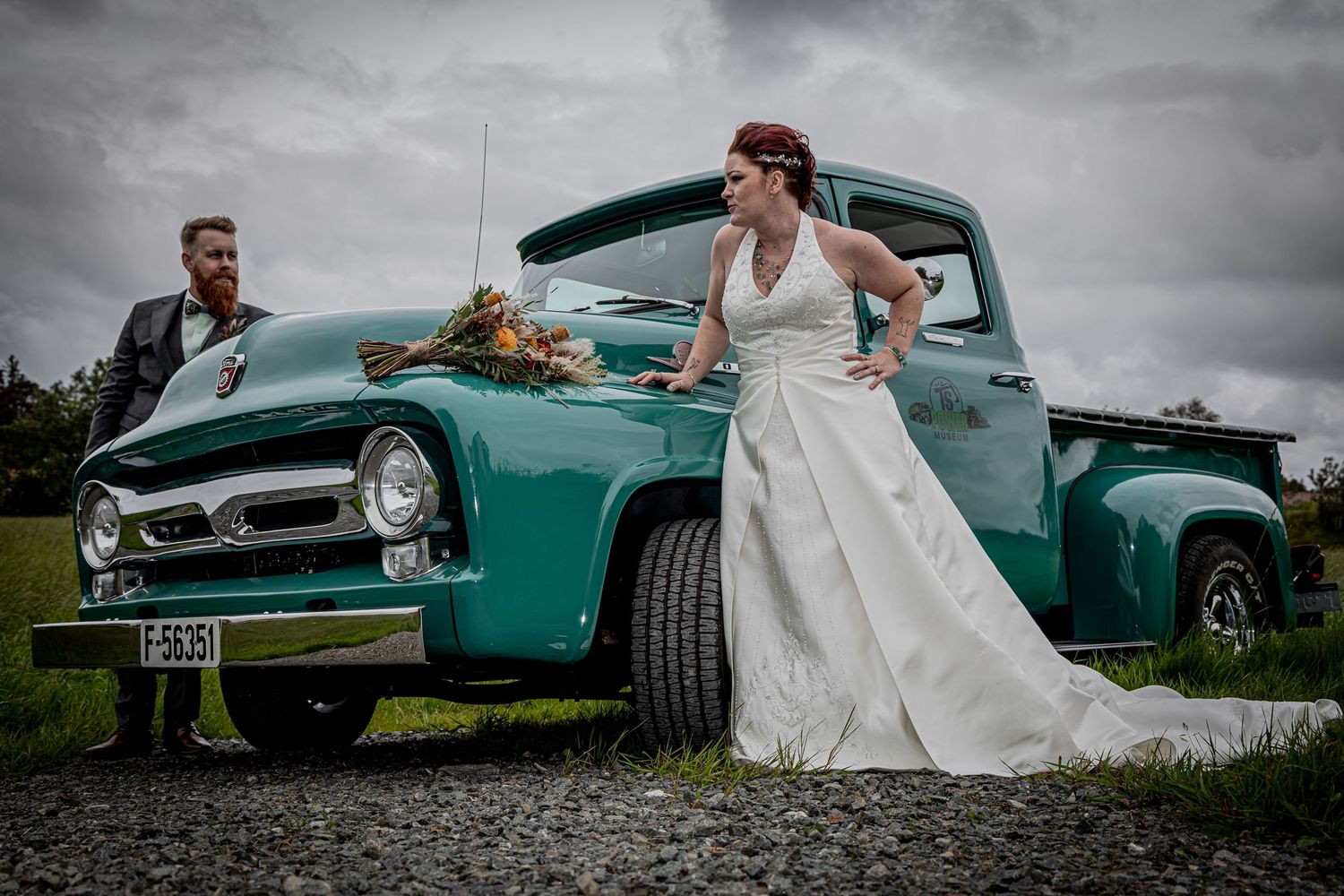 Bride in white gown leans against vintage teal Ford pickup truck with groom beside it on cloudy day.
