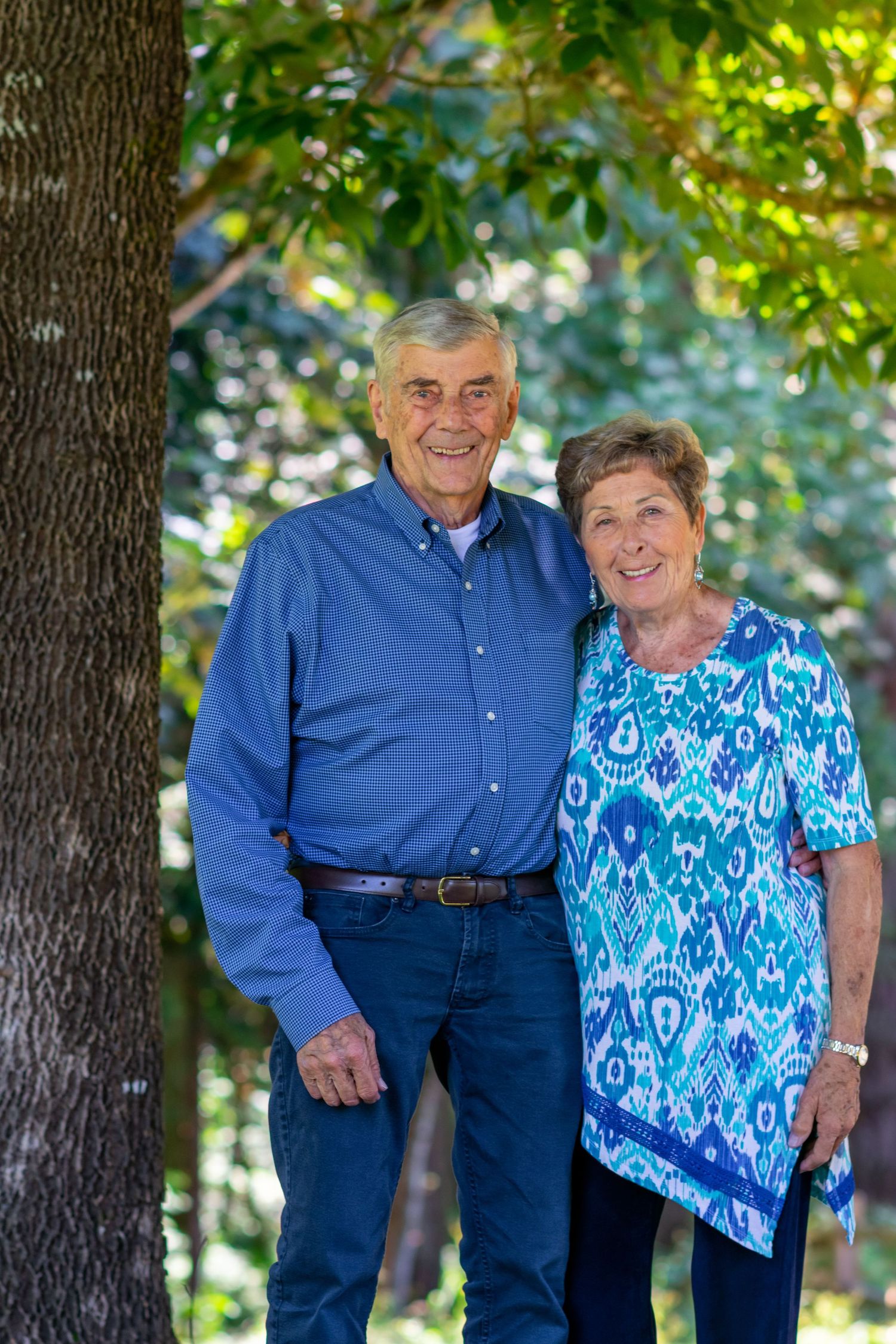 Older couple wearing complimentary outfits standing next to a tree and smiling at camera