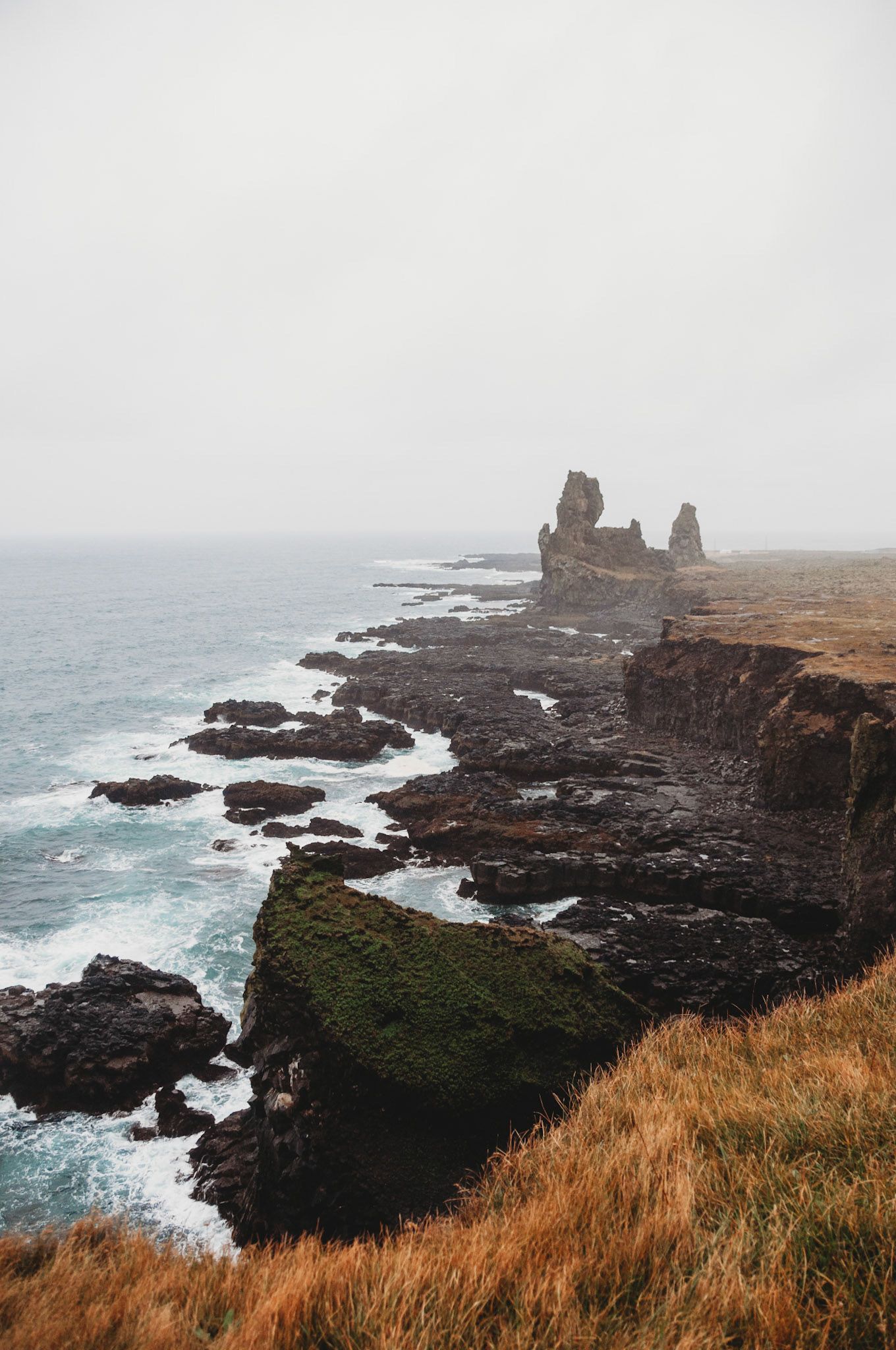 Dramatic rocky coastline with jagged cliffs and crashing waves against dark volcanic rocks in misty weather.