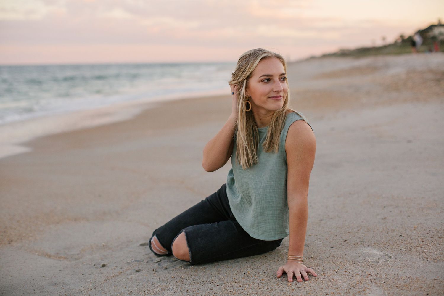 Young woman in teal tank top sitting on beach at sunset.
