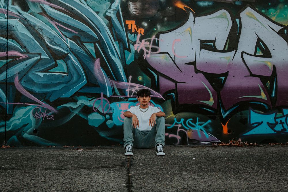 Young man in a white t-shirt and jeans sits on the ground in front of a colorful graffiti mural.