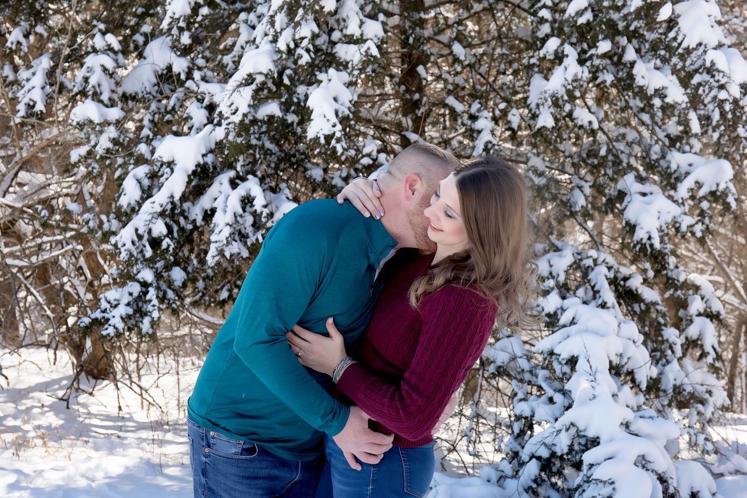 Couple in burgundy and teal sweaters share romantic moment in snowy woods.