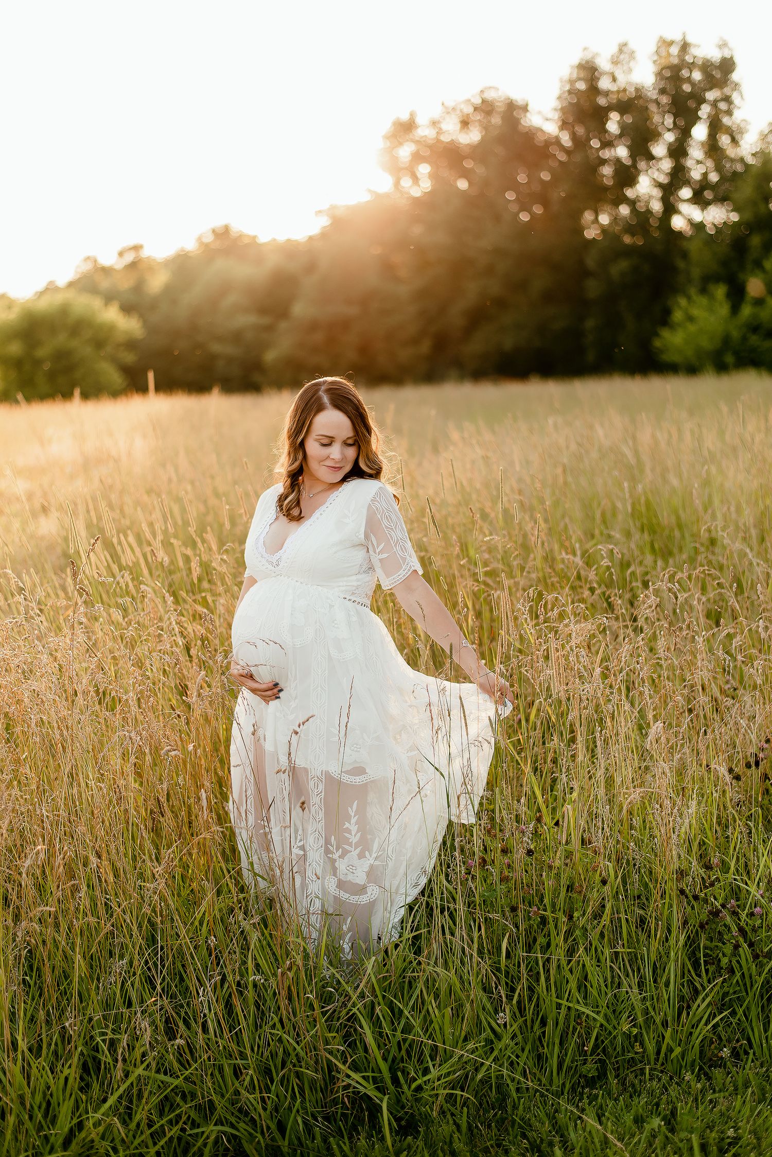 Pregnant woman in white lace dress walking through tall golden grass field at sunset.
