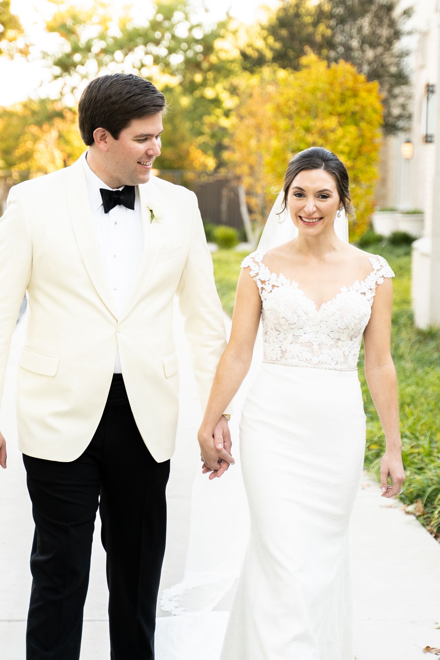 Wedding couple walking together outdoors in formal white attire against autumn foliage.