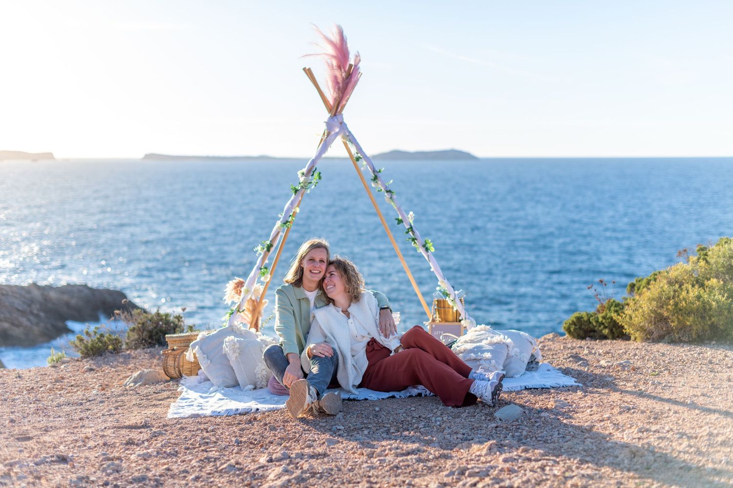 A lesbian couple sit under a teepee structure in Ibiza at the top of a cliff with view of a blue sea