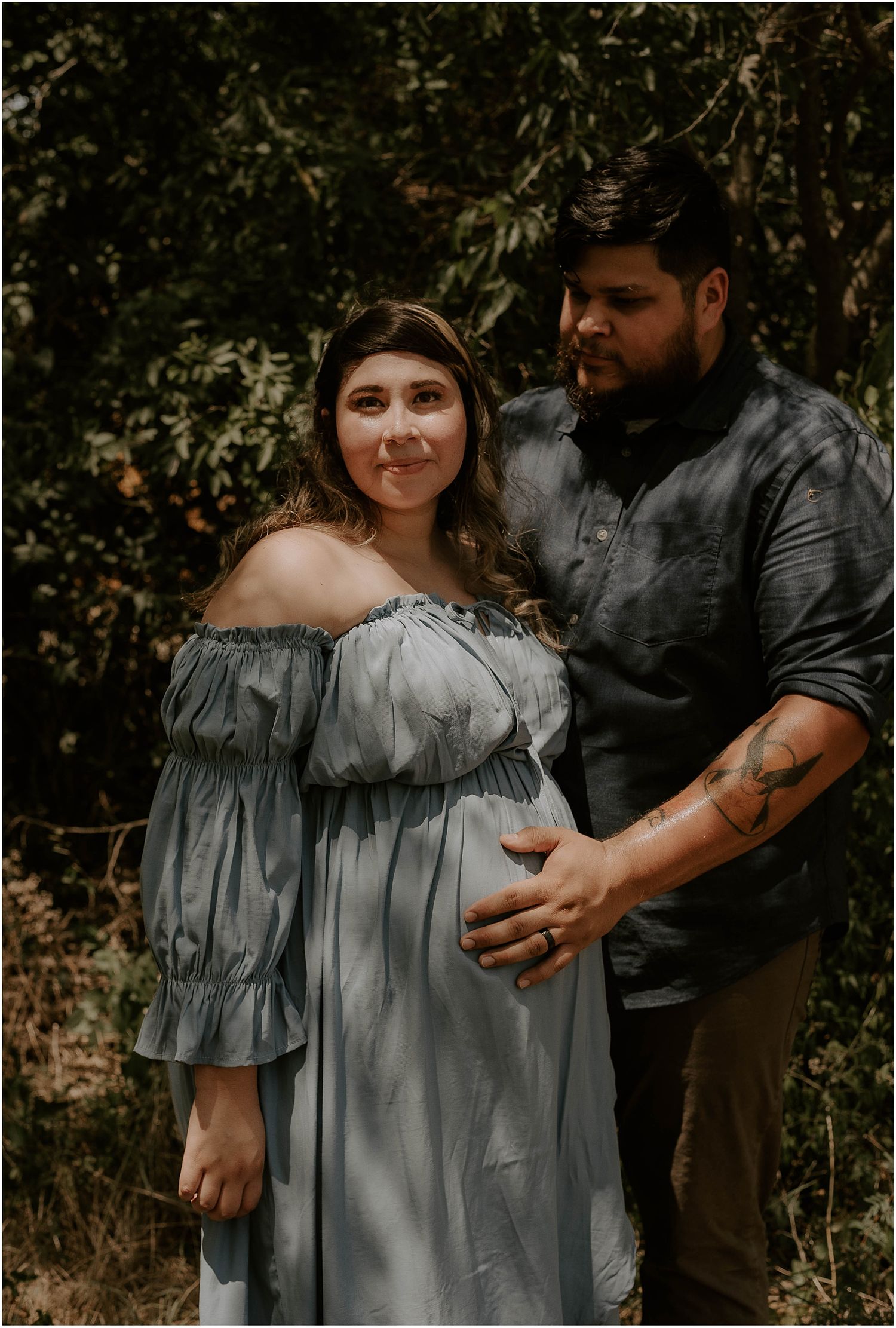Rustic outdoor maternity portrait featuring couple in blue off-shoulder dress and dark shirt.