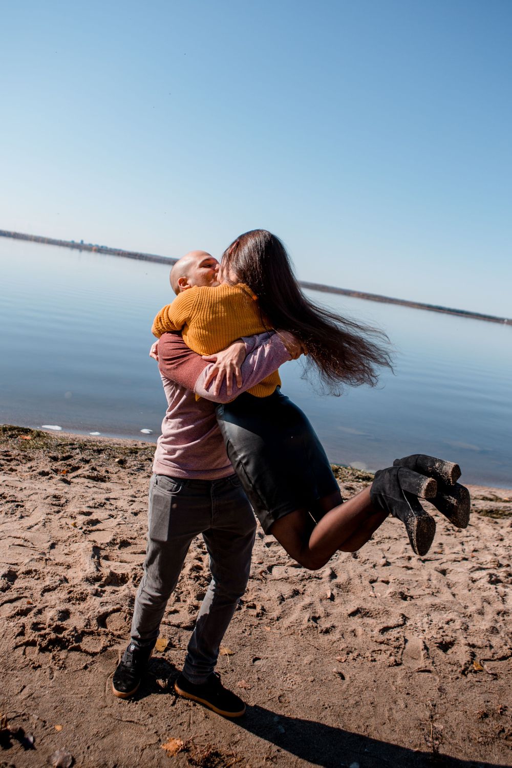 Playful couple spinning on beach with clear blue sky and water in background.