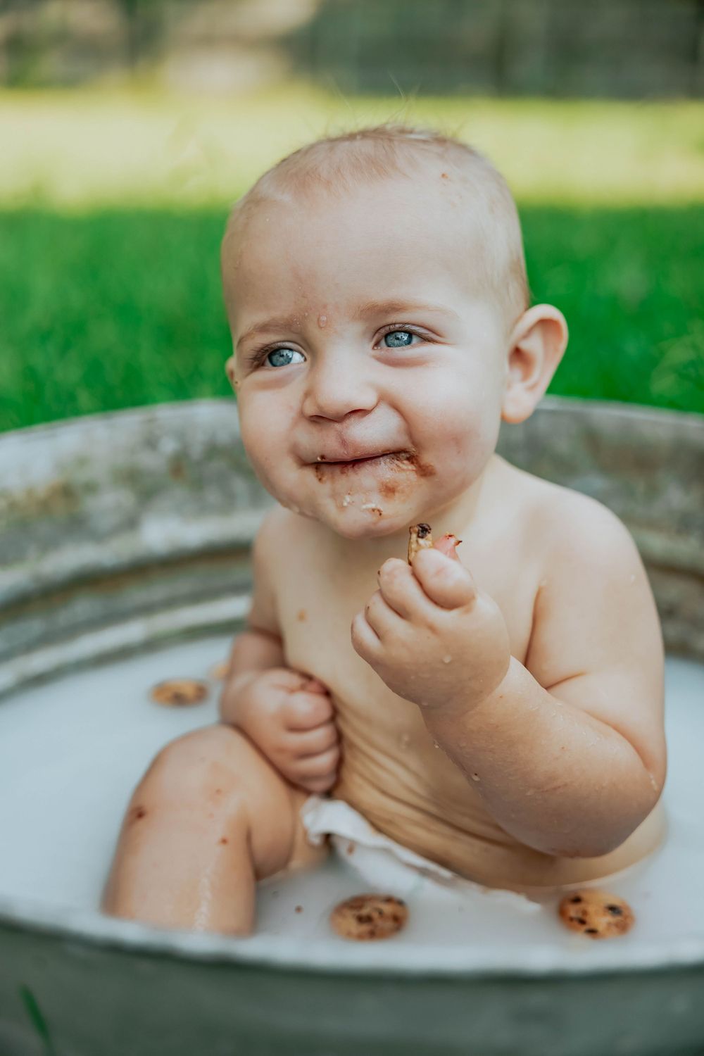 A messy baby smiles while playing in a white basin outdoors.
