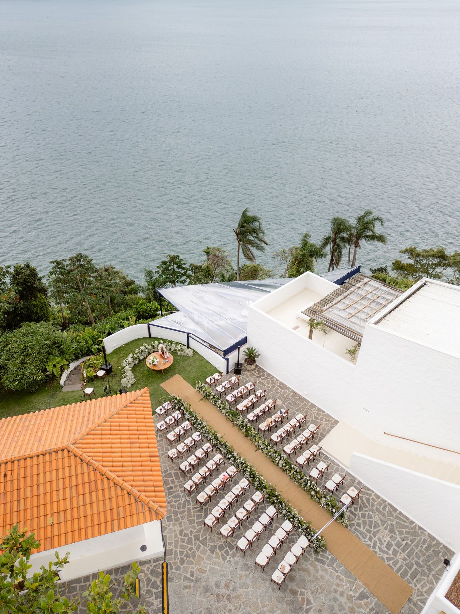 Aerial view of a coastal wedding venue with white chairs set up for an outdoor ceremony overlooking the ocean.