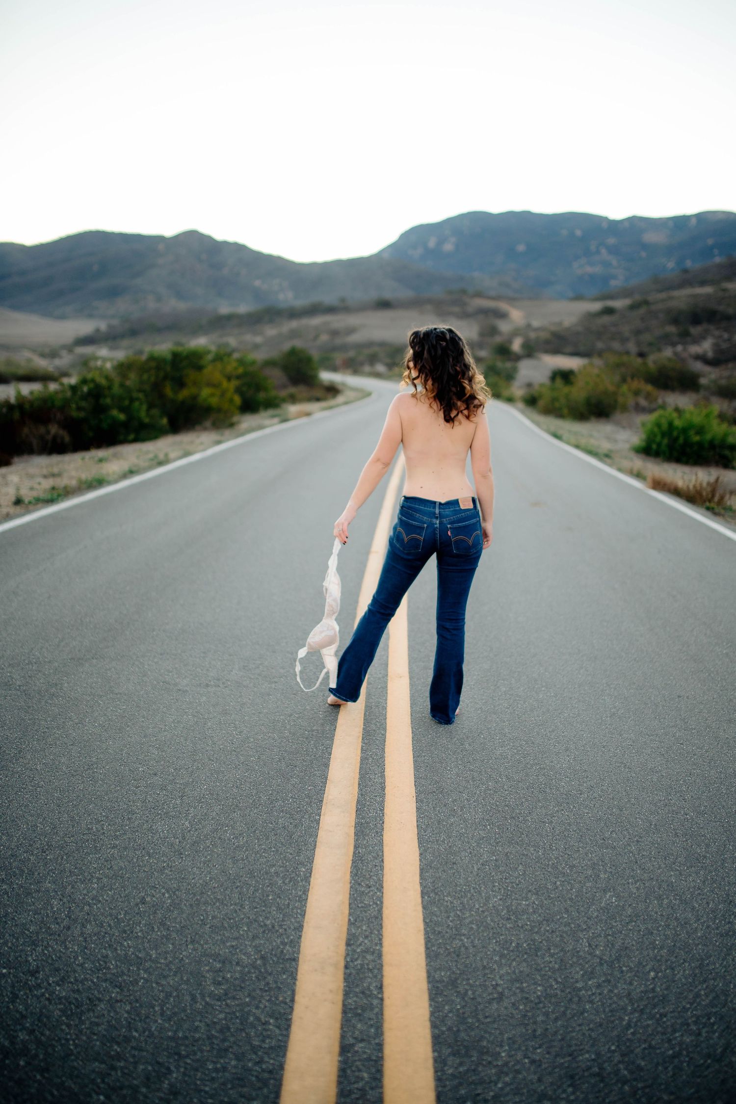 Women in the middle of a winding rode in jeans, topless, holding a bra to her left as she walks away from the camera