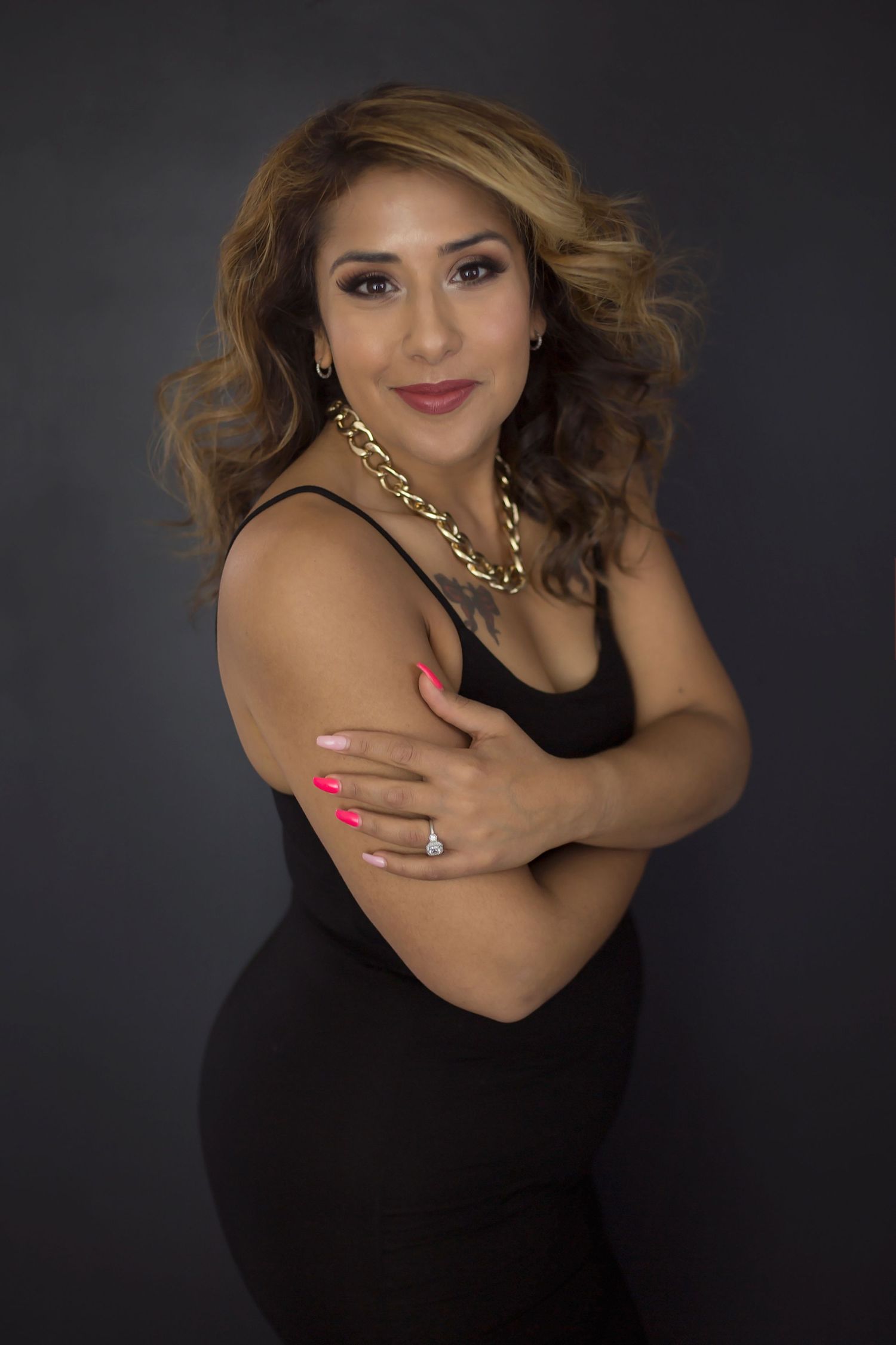 A woman in a black dress with a gold chain necklace and pink nails poses against a dark background.