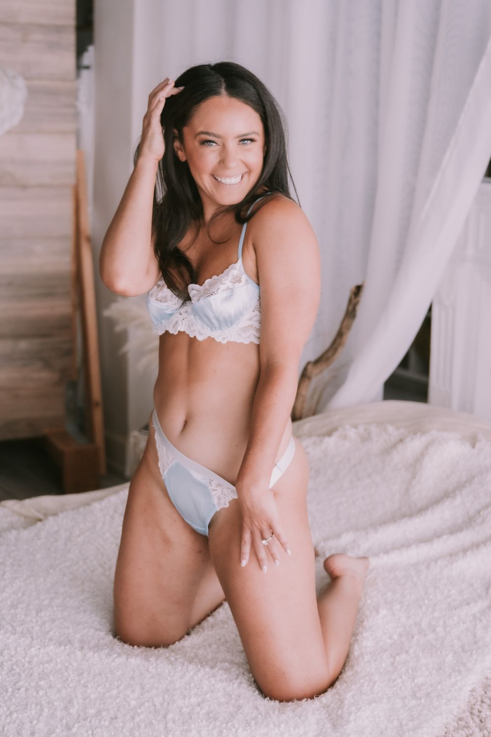 Smiling brunette woman in white lace lingerie kneeling on a fluffy white bed in a boudoir setting.