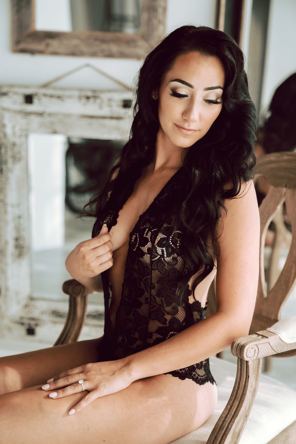 Brunette woman in black lace bodysuit sitting gracefully on a chair in an elegant room.