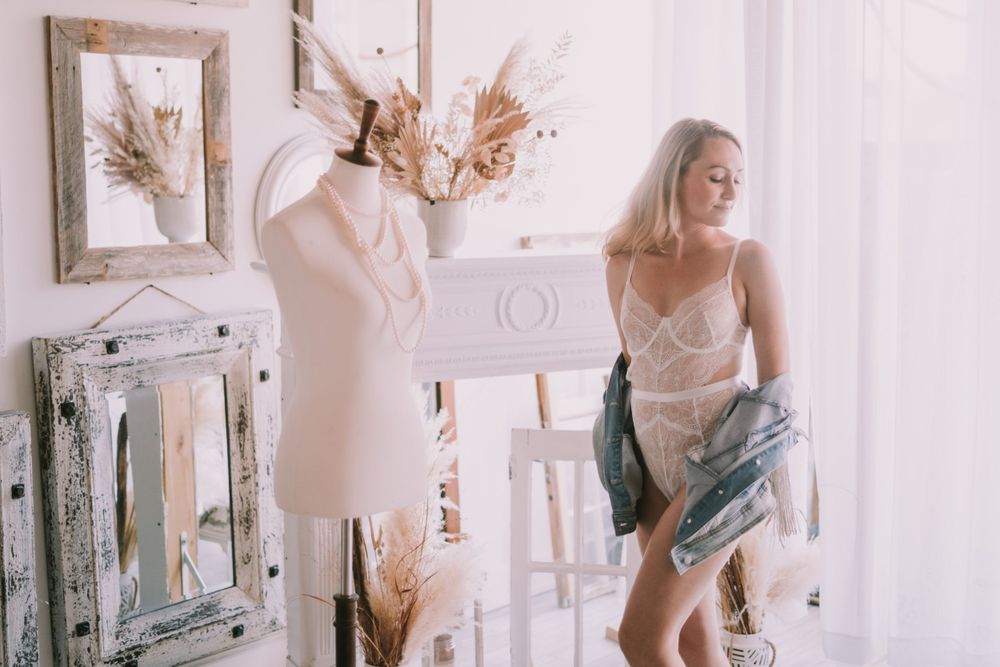 Elegant blonde woman in sheer lingerie removing a denim jacket in a bright white vintage-styled boudoir studio.