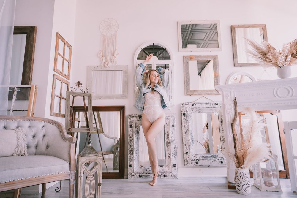 Blonde woman in a bodysuit posing among vintage mirrors and elegant decor in a bright white boudoir studio.