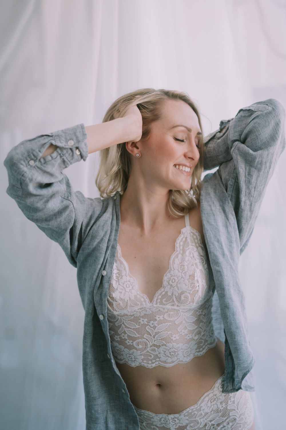 Smiling blonde woman in white lace bralette and grey shirt posing with hands in hair against sheer curtains.