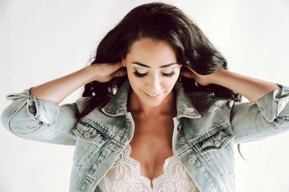 Smiling brunette woman in denim jacket and white lace top with hands in her hair.