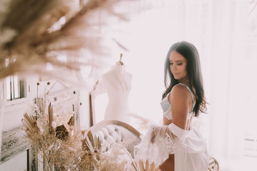 Brunette woman in white lingerie and robe examining a dress form mannequin in a softly lit studio.