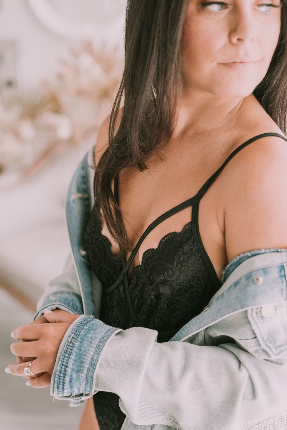Close-up portrait of brunette woman in black lace bralette and denim jacket with a confident expression.