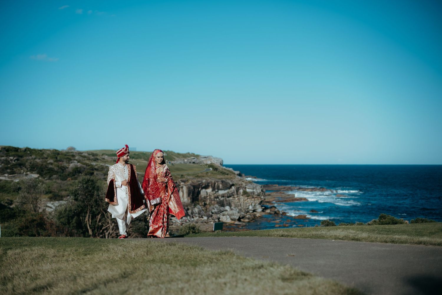 Two people walk along a scenic coastal path with rugged cliffs and blue ocean views on a bright sunny day.