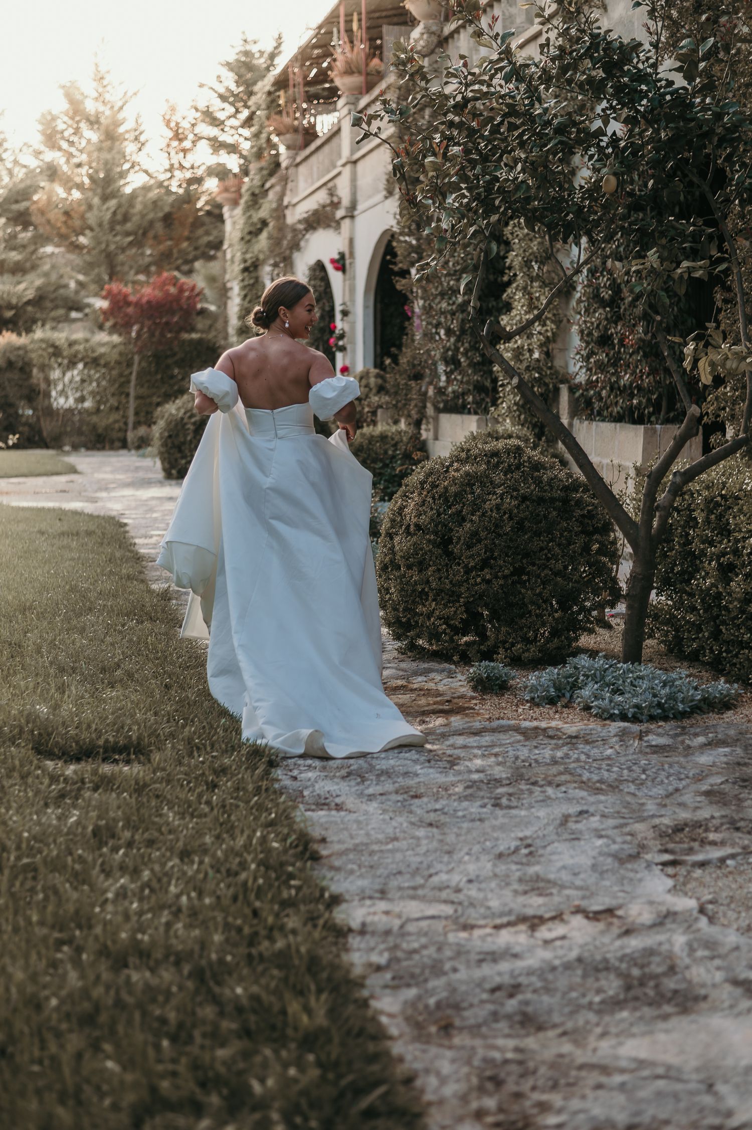 Elegant bride in off-shoulder white gown walking along stone path beside historic building at dusk.