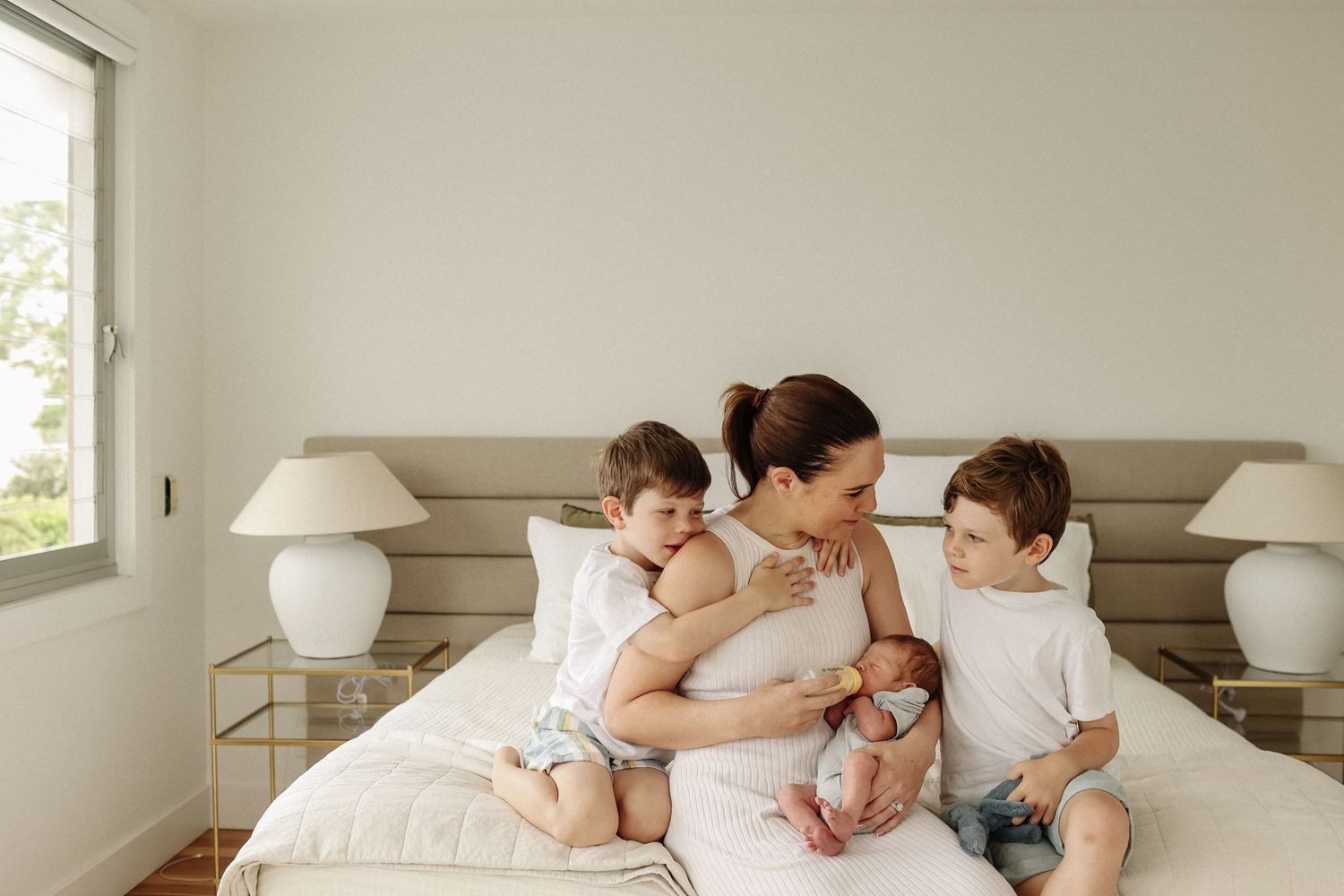 Mother sitting on bed with newborn baby and two young boys in a bright minimalist bedroom.