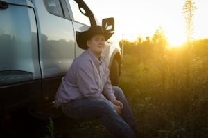 A person in a cowboy hat sits against a truck during golden hour sunset.