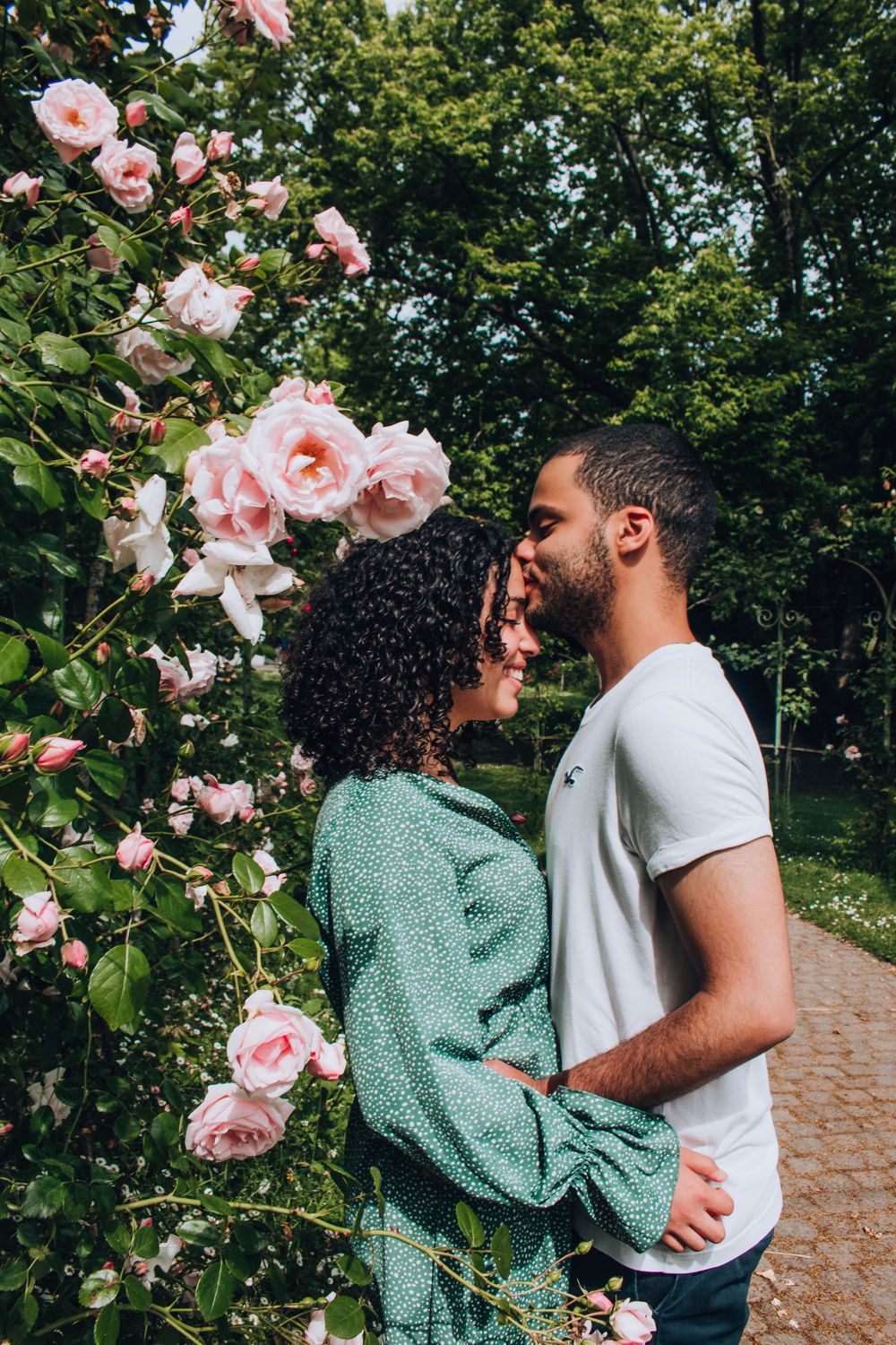 A couple standing close together beside a blooming pink rose bush in a garden.
