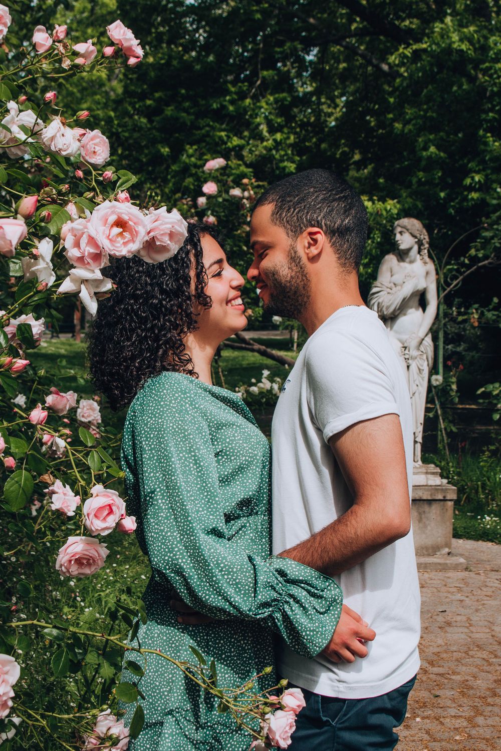 A couple embracing in a garden with pink roses and a classical statue.