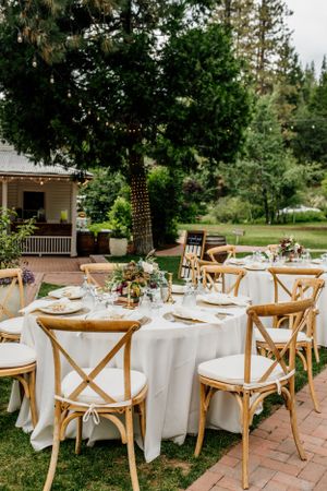 Elegant outdoor wedding reception setup with white tablecloths and wooden cross-back chairs.