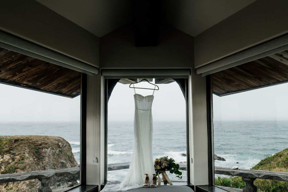 White wedding dress hanging in window frame with ocean view and rocky coastline.