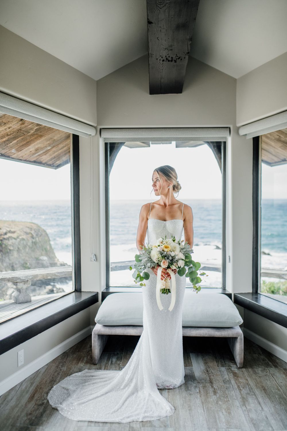 Bride in elegant white gown stands in coastal room with panoramic ocean views.