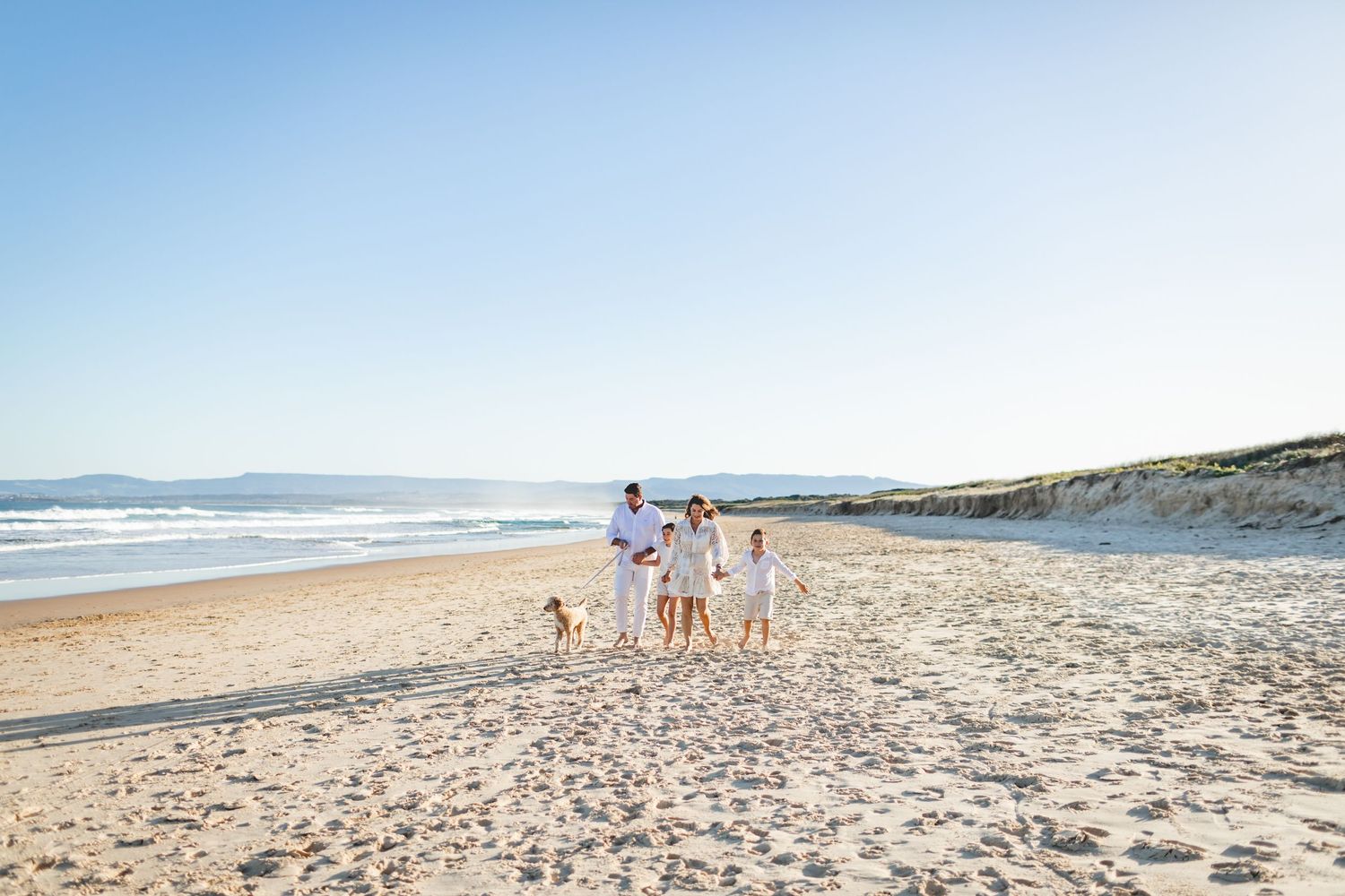 Family and dog walking along a scenic beach shoreline on a sunny day.
